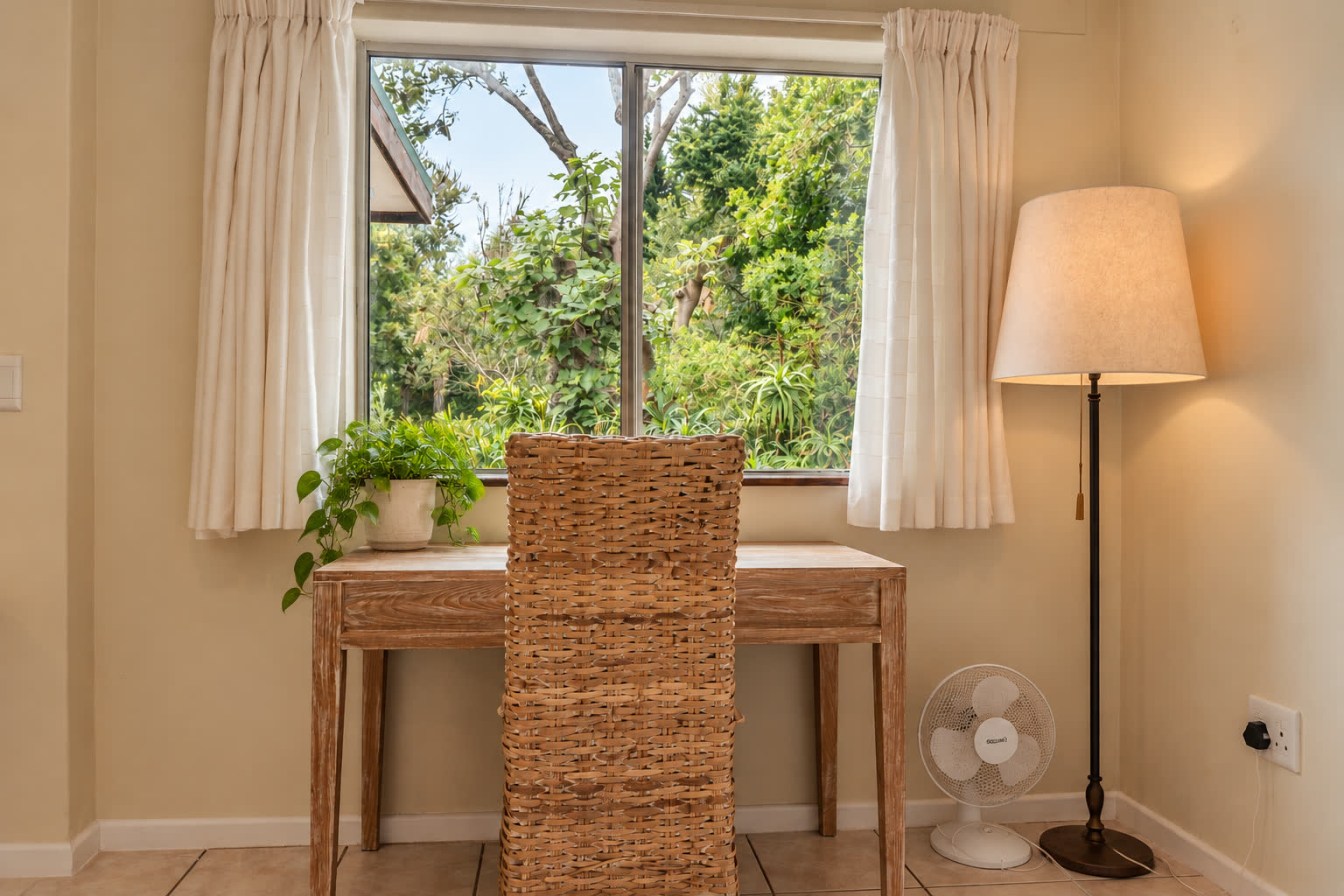 Study nook at Loerie Chalet with a light wood desk and woven chair facing a window onto the garden, a floor lamp and small fan beside it