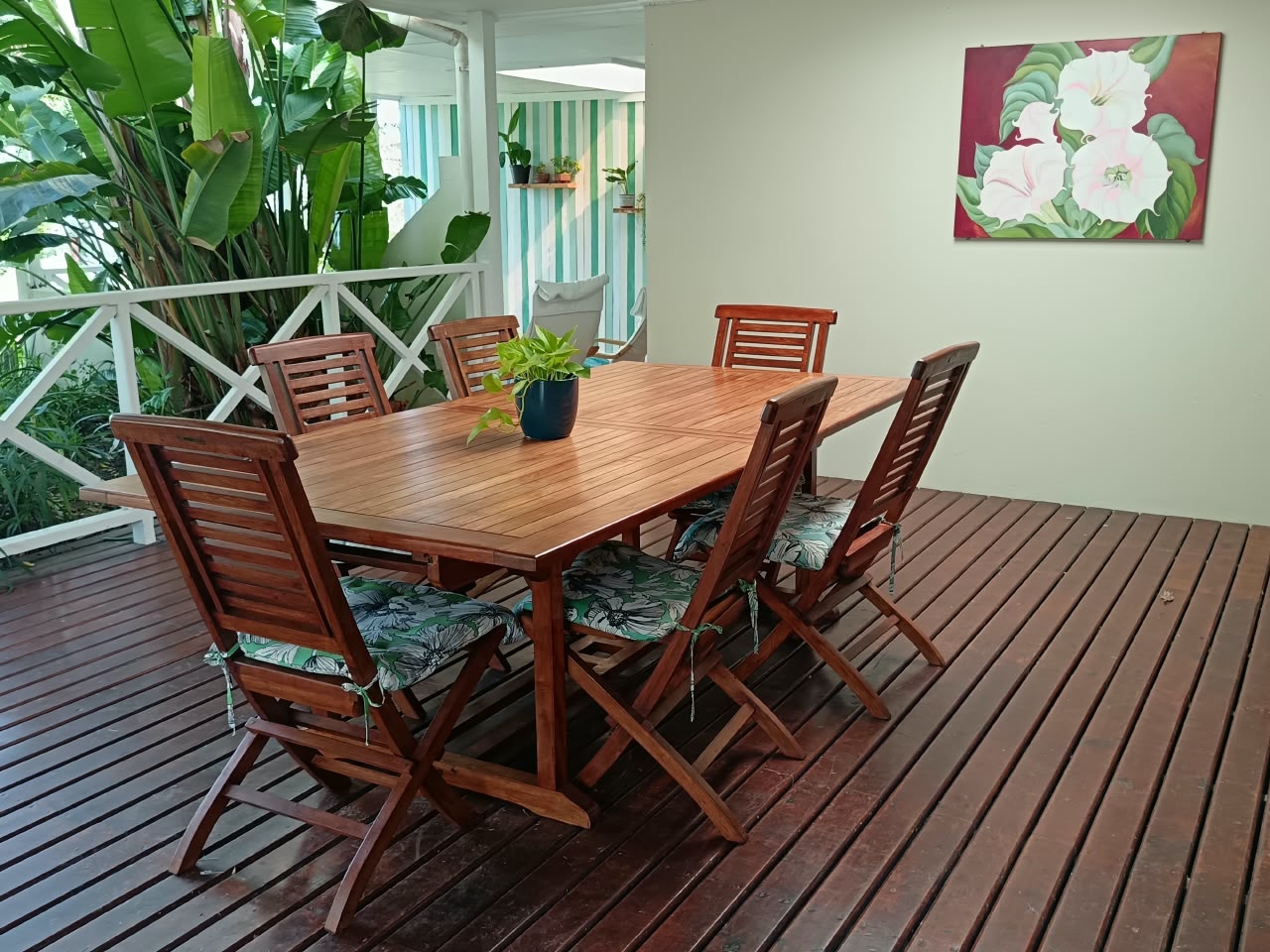 Covered wooden deck at Fish Eagle Chalet with a slatted dining table, six chairs with botanical cushions and a white railing among tropical plants