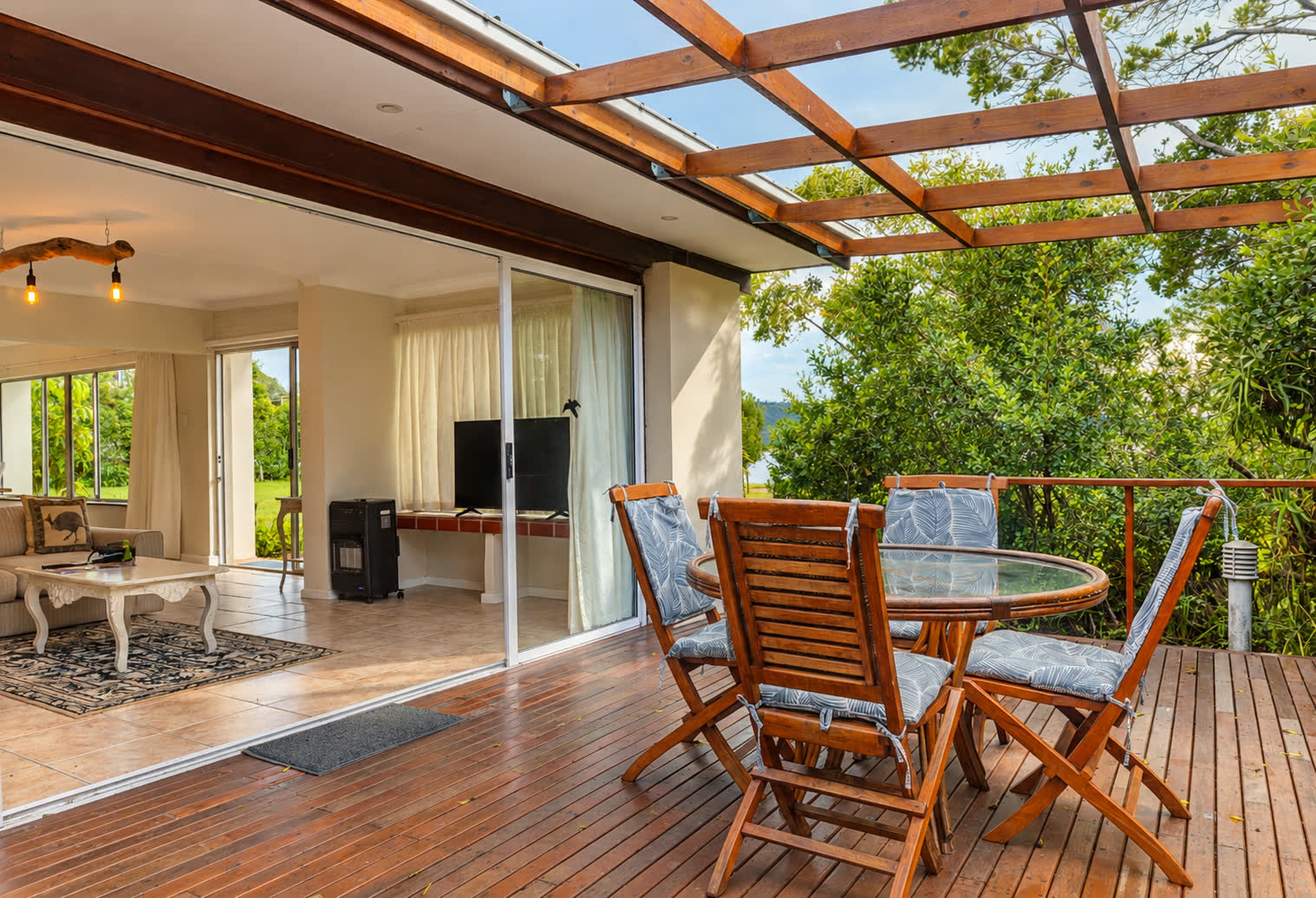 Loerie Chalet’s timber deck under an open pergola with a round wooden dining table and four cushioned chairs, sliding doors open to the living room